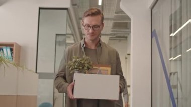 Yeni bir iş bulduktan sonra elinde kağıt kutuyla modern ofis koridorunda yürüyen ve akıllı telefondan konuşan genç ve yetişkin bir adamın orta yavaş çekimi.