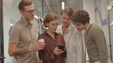 Kahve molası sırasında ofis koridorunda duran akıllı telefondan bir şeyi tartışan dört erkek ve kadından oluşan genç yaratıcı ekibin orta yavaş çekimi.