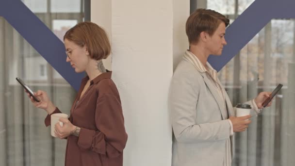 两名女同事背对背站在柱子旁 在办公室喝咖啡休息时在智能手机上打滚 然后开始交谈的慢镜头 — 图库视频影像