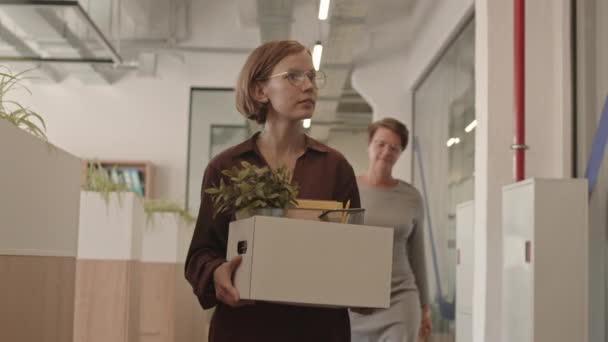 女性上司と話している新しいオフィスに沿って歩いて彼女のものと紙の箱を運ぶ若い陽気な女性の中低速ショットを追跡します 従業員としての女性の最初の日 — ストック動画