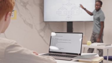 Sınıfta masada oturan, dizüstü bilgisayarda kod yazan, bulanık öğretmenin konferansını dinleyen çocuğun omuz üstü görüntüsü.