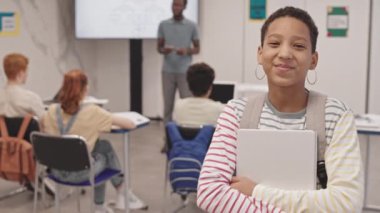 Sırt çantası takan, dizüstü bilgisayarı olan, ders sırasında sınıfta duran, konuşan, gülümseyen ve kameraya bakan neşeli siyahi bir kız.