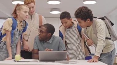Dört farklı ortaokul öğrencisinden oluşan orta sınıf öğrencileri sınıftaki masasında oturan, dizüstü bilgisayar kullanan ve konuşan genç siyahi erkek öğretmenlerle konuşuyorlar.