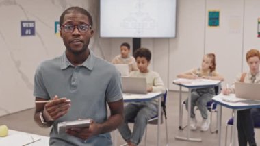 Orta yaşlı siyah erkek öğretmen gözlük takıyor, elinde kalem ve defter var. Ortaokuldaki bulanık sınıfın önünde dikilip kameraya bakıyor.