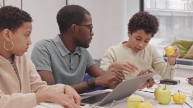 Orta uzunlukta genç siyahi erkek öğretmen genç kız ve melez oğlanla aynı masada oturuyor, elmalar ve limonlarla elektriği ileten deneyler yapıyor.