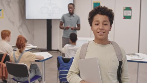 一个快乐的小孩背着背包 拿着手提电脑 上课的时候站在教室里 微笑着 看着镜头 — 图库视频影像