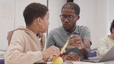 Genç siyahi öğretmen ve ortaokul öğrencisi sınıftaki masada oturmuş elektrik ve limonla deney yapıyor, konuşuyor.