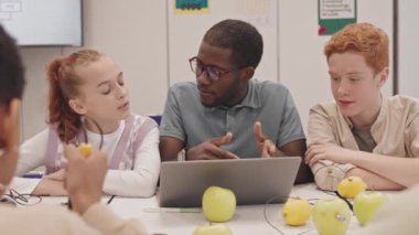 Orta boy erkek fizik öğretmeni gözlüklü, dizüstü bilgisayarlı, elmalı ve limonlu masada oturan, öğrencilerle konuşan
