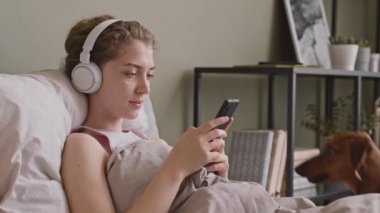 Haftasonu tembel bir şekilde yatakta dinlenirken, 20 'li yaşlarının başında, kulaklıkla müzik dinleyen ve tatlı daksund köpeğiyle oynayan mutlu genç bir kadının belden aşağı çekimini yapın.