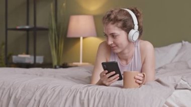 Ev elbiseleri ve kulaklıklar takmış, sabah kahvesinin tadını çıkarırken arka planda sevimli bir köpekle oynayan rahat genç bir bayanla orta boy yakın çekim.