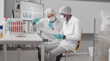 Yüz maskesi, eldiven, tıbbi önlük ve şapka takan iki erkek bilim adamının beliyle laboratuvar masasında oturmuş test tüplerini inceliyor, konuşuyor ve belgeleri okuyorlar.