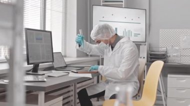 Orta uzunlukta, yüz maskesi, eldiven, tıbbi önlük ve şapka takan bir bilim adamı laboratuarda oturmuş test tüpündeki sıvıyı inceliyor, arkasını dönüyor ve iş arkadaşıyla konuşuyor.