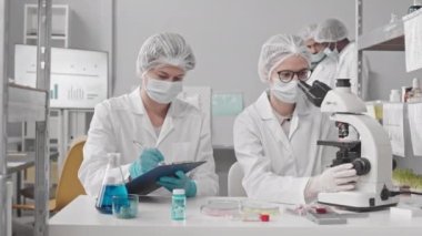 Yüz maskeleri, eldivenler, tıbbi elbiseler ve şapkalar giyen iki kadın bilim adamının mikroskop kullanarak, konuşarak ve notlar alarak laboratuvarda masasında oturması, arka planda yürüyen erkek meslektaşları.