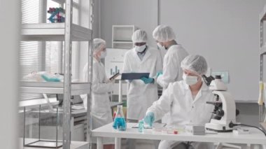 Orta uzunlukta, yüz maskesi, eldiven, tıbbi önlük ve şapka takan, mikroskop kullanan, arka plan üzerinde çalışan çeşitli meslektaşları olan beyaz bir kadın.