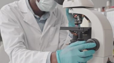 Tıbbi üniforma giyen, laboratuarda mikroskop kullanan genç Afrika kökenli Amerikalı bilim adamının...