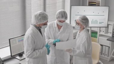 Çift ırklı erkek ve beyaz kadın bilim adamlarından oluşan yüksek açılı bir ortam. Çalışma üniforması giyen iki kadın gündüz vakti laboratuvarda oturmuş sohbet ediyorlar.