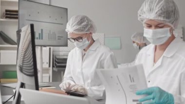 Tıbbi elbiseler, maskeler, şapkalar ve eldivenler giyen iki zeki beyaz kadın gündüzleri laboratuarda masasında oturuyor ve konuşuyor.