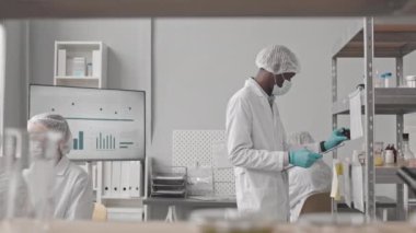 Orta uzunluktaki Afrikalı Amerikalı adam, laboratuvarda raflarda duruyor, not alıyor, erkek çift ırklı meslektaşı kalkıp bilim adamıyla konuşuyor.