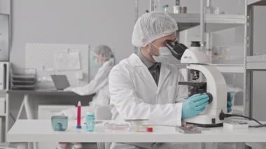 Tıbbi üniforma giyen, laboratuarda masasında oturan, mikroskop kullanan ve not alan genç erkek çift ırklı bilim adamı.