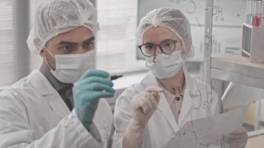 Laboratuvarda cam duvara yapısal kimyasal formüller yazan melez bir adam, yanında kağıt tutan beyaz bir meslektaşı, tıbbi giysiler ve maskeler giyen bilim adamları.