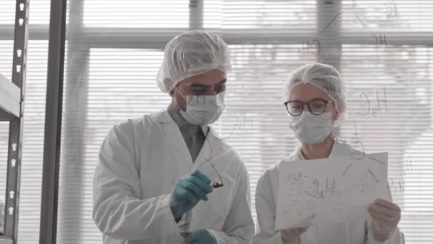 研究室のガラスの壁に構造化学式を書いている獣医師の腰のアップ 近くに立って紙を保持する女性白人同僚 医学のガウンとマスクを身に着けている科学者 — ストック動画