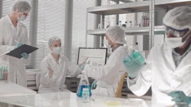 Medikal giysiler, maskeler ve eldivenler giyen, gündüzleri laboratuvarda çalışan ve konuşan orta büyüklükte çok ırklı bilim adamları.