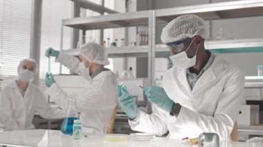 Tıbbi önlük ve gözlük takan, elinde petri kabı tutan, forseps ile biyolojik materyal alan ve yoğun bir laboratuvarda masa başında oturan Afrika kökenli Amerikalı erkek bilim adamı.