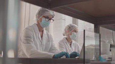 Tıbbi önlük, maske ve şapka takan, iş günü masa başında bilgisayar kullanan ve konuşan iki beyaz kadın laboratuvar asistanının düşük açılı görüntüsü.