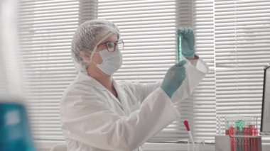 Tıbbi önlük ve maske takan, gündüzleri araştırma merkezinde masasında oturan, test tüplerinde kimyasallarla çalışan zeki, beyaz bir kadının düşük açılı görüntüsü.