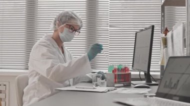 Tıbbi önlük ve maske takan, gündüzleri araştırma merkezinde masasında oturan, test tüplerinde kimyasallarla çalışan zeki, beyaz bir kadın.