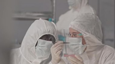 Tehlikeli madde önlüğü, koruyucu gözlük ve maskeler takan, araştırma merkezinde oturan, Petri kabındaki biyolojik materyalleri analiz eden Afrikalı Amerikalı ve beyaz erkek laboratuvar çalışanlarının omuz hizasında.