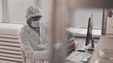 Tıbbi önlük, gözlük ve maske takan, gündüzleri araştırma merkezinde masasında oturan test tüplerindeki kimyasallarla çalışan kıyılmış iş arkadaşı