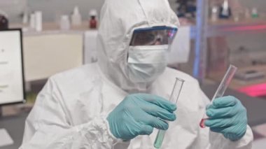 Tehlikeli madde önlüğü, gözlük ve maske takan, araştırma merkezinde duran, test tüplerinde kimyasalları karıştıran Afrikalı Amerikalı erkek laboratuvar çalışanı yüksek açılı göğüs röntgeni.