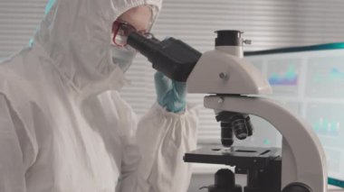 Tehlikeli madde giysisi, eldiven, gözlük ve tıbbi maske giyen kadın bilim adamlarının göğüs göğsü. Araştırma merkezinde, mikroskop kullanarak oturuyorlar.