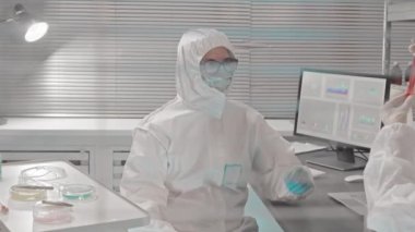 Tehlikeli madde giysileri, eldivenler ve tıbbi maskeler giyen iki bilim adamının test tüplerindeki kimyasallarla birlikte çalışma masasında oturduğu cam duvarın ortası.
