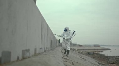 Erkek ekolojistin koruyucu giysisi ve botlarıyla kirli bölgede ilerlerken özel bir sıvıyı betona püskürtürken yavaş çekimleri.