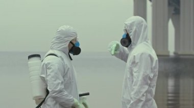 Kimyasal kostümlü ve solunum maskeli iki farklı bilim adamının orta yavaş portresi çevresel felaketin yaşandığı bölgede biyolojik tehlikelerin ortadan kaldırılmasını tartışıyor.