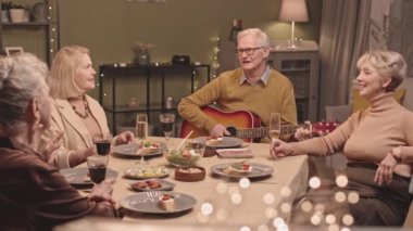 Gitar çalan ve şarkı söyleyen yaşlı bir adamın kadın arkadaşlarıyla yemek masasında oturup birlikte iyi vakit geçirdiği orta derece yavaş bir çekim.