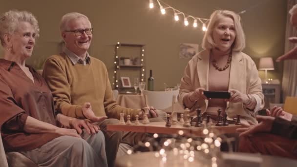 明るい高齢者のグループの中低速ショット一緒に自宅で夜を過ごす チェスや話をする 良いゲームの後に手を振る男性と女性の一人は スマートフォンでビデオを撮る — ストック動画