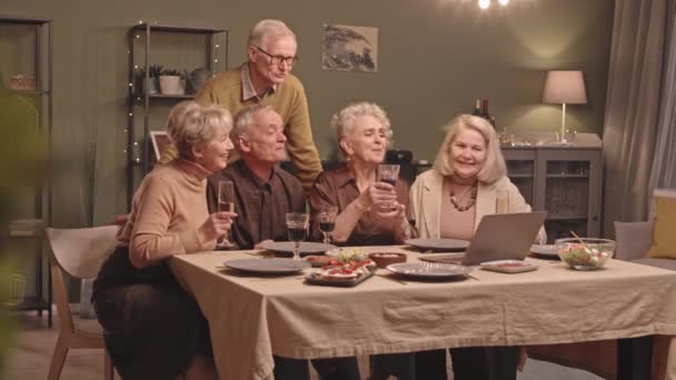 中低速ショットの幸せなシニア男性と女性持っていますビデオチャット上のラップトップでお祝いのディナーテーブル クリンクガラスと支出楽しい時間一緒に — ストック動画