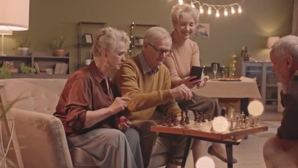 夕方の照明付きの大きな居心地の良いリビングルームでチェスをしながら楽しんでいる高齢者のグループの中のショット スマートフォンでゲームを録画して笑っている女性の一人 — ストック動画