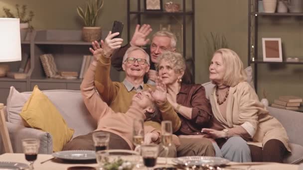 中低速ショットの5幸せな白人高齢者一緒にソファの上に居心地の良いリビングルームで休憩し 友人とスマートフォンでビデオ通話を持ちます 手を振って カメラで話して — ストック動画