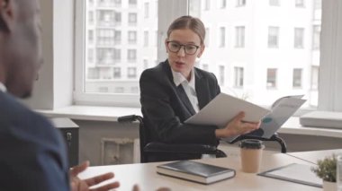 Tekerlekli sandalyedeki beyaz kadın CEO 'nun elinde belgelerle gündüz vakti konferans masasında oturan kırpılmış çalışanla konuştuğu bir ortam.