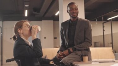 Orta uzunlukta, gri takım elbiseli siyah bir adam ve tekerlekli sandalyedeki kısa saçlı beyaz kadın modern ofisteki masada oturuyor, konuşuyor ve kahve içiyor.