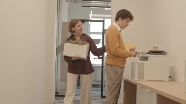 オフィスのコピー機の隣に立つ職場への方向性を同僚に求めるものと彼女のものと紙箱を運ぶ新しい女性従業員の中低速ショット — ストック動画