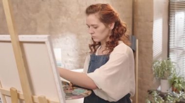 Kıvırcık, kızıl saçlı, önlük giyen, gündüz vakti resim stüdyosunda fırçayla resim yapan beyaz bir kadın.