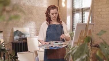 Genç kızıl saçlı beyaz kadın önlük giyiyor, gündüz resim stüdyosunda resim yapıyor.