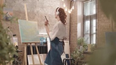 Orta uzunlukta, kulaklık takan, müzik eşliğinde dans eden, stüdyo tuvalinde boya fırçasıyla soyut sanat eserleri çizen kızıl saçlı bir kadın.