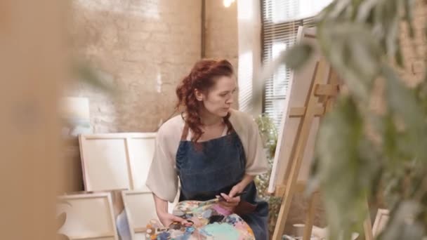 앞치마를 스튜디오에 들어가 앉아서 캔버스에 그림을 그리는 여성을 추적하는 — 비디오