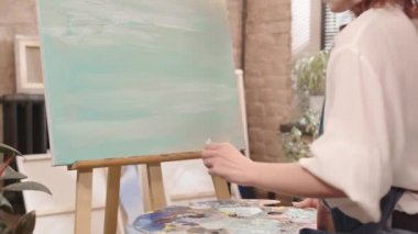 Yan görüntüde, önlük takan, gündüz vakti sanat stüdyosunda macun bıçağıyla soyut sanat eserleri çizen kızıl saçlı beyaz kadın görülüyor.